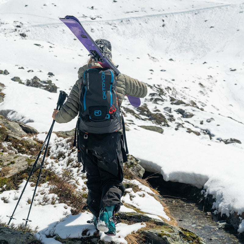 Women walking by snowy river carrying Faction Agent 2X purple skis over her shoulder.
