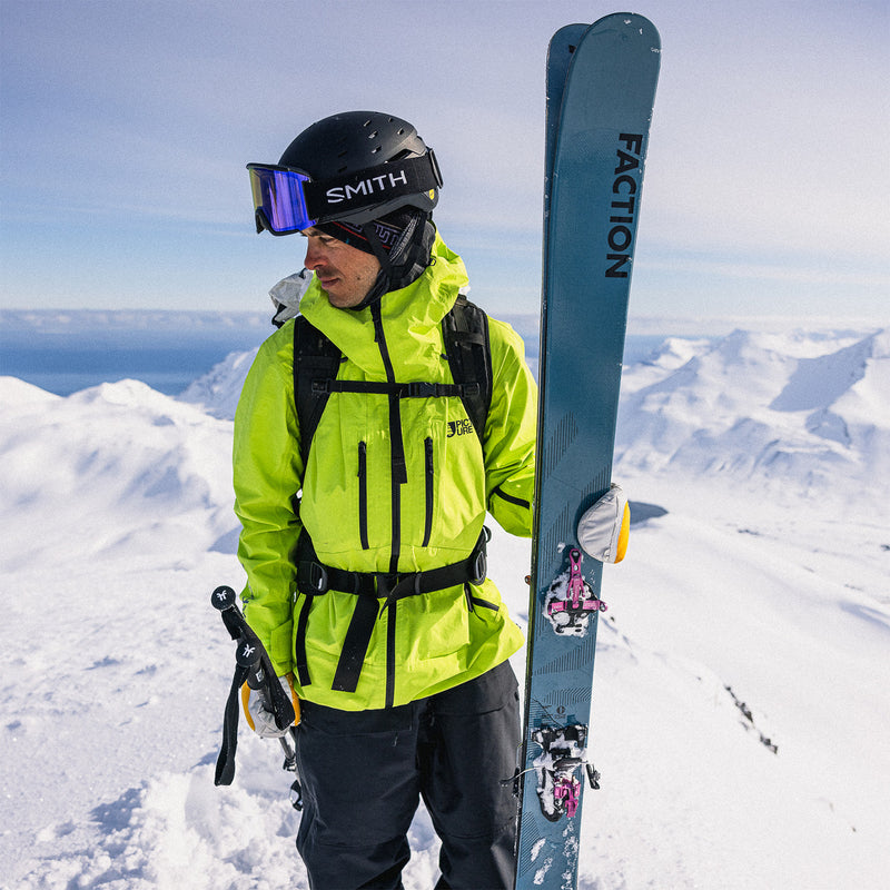 Man in bright yellow jacket and black helmet holding a blue ski on a mountain top.