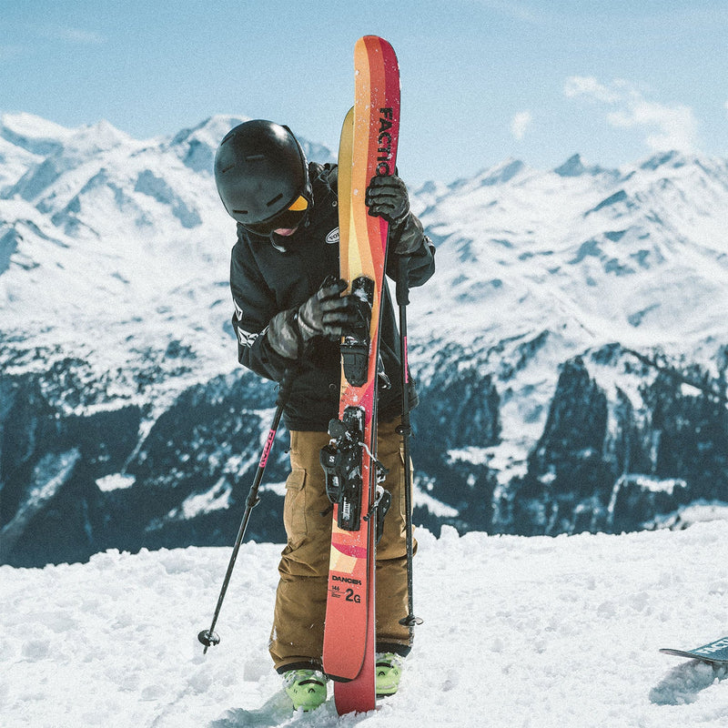 Young skier holding Faction Dancer Grom red skis in a snowy alpine setting. 2026