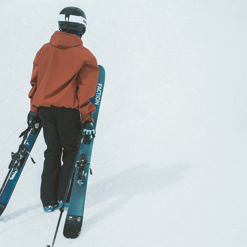 Young skier in an orange jacket wearing a helmet walking up a snow park carrying the Faction Studio Grom skis in his hands.