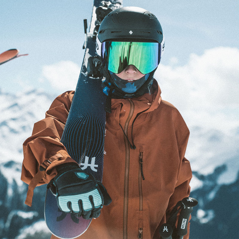 Young skier in an orange jacket wearing a helmet and green ski goggles carrying the Faction Studio Grom over his shoulder.