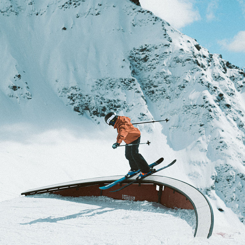 Young skier in an orange jacket and wearing a helmet doing a snowpark rail with Faction Studio Grom skis.