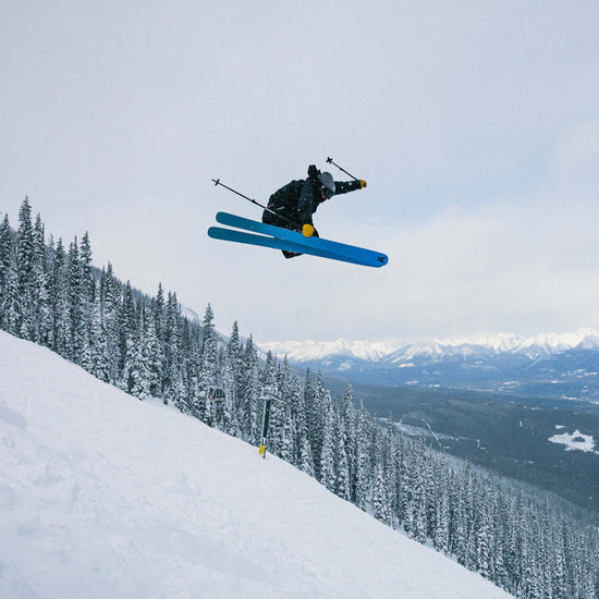 Skier in mid-air showing the blue base of the Faction Dancer 106 Ti against a snowy mountain landscape with trees