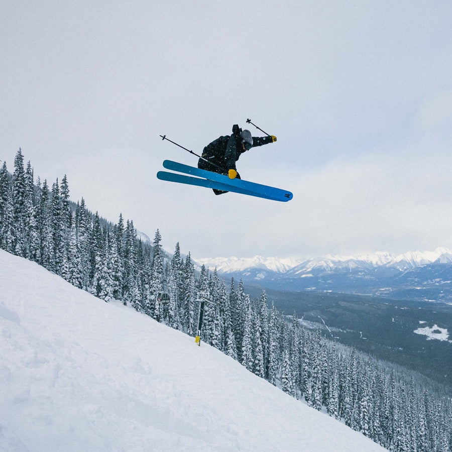 Skier in mid-air showing the blue base of the Faction Dancer 106 Ti against a snowy mountain landscape with trees