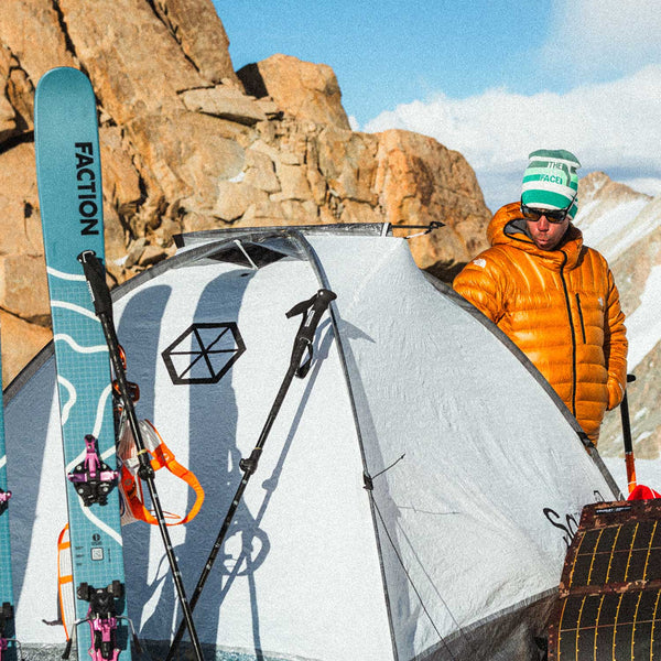 Person standing next to a tent with a skier's backpack and a ski with 'Faction' branding in a mountainous area.