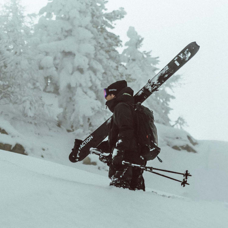 Person carrying skis and poles through a snowy landscape with trees.